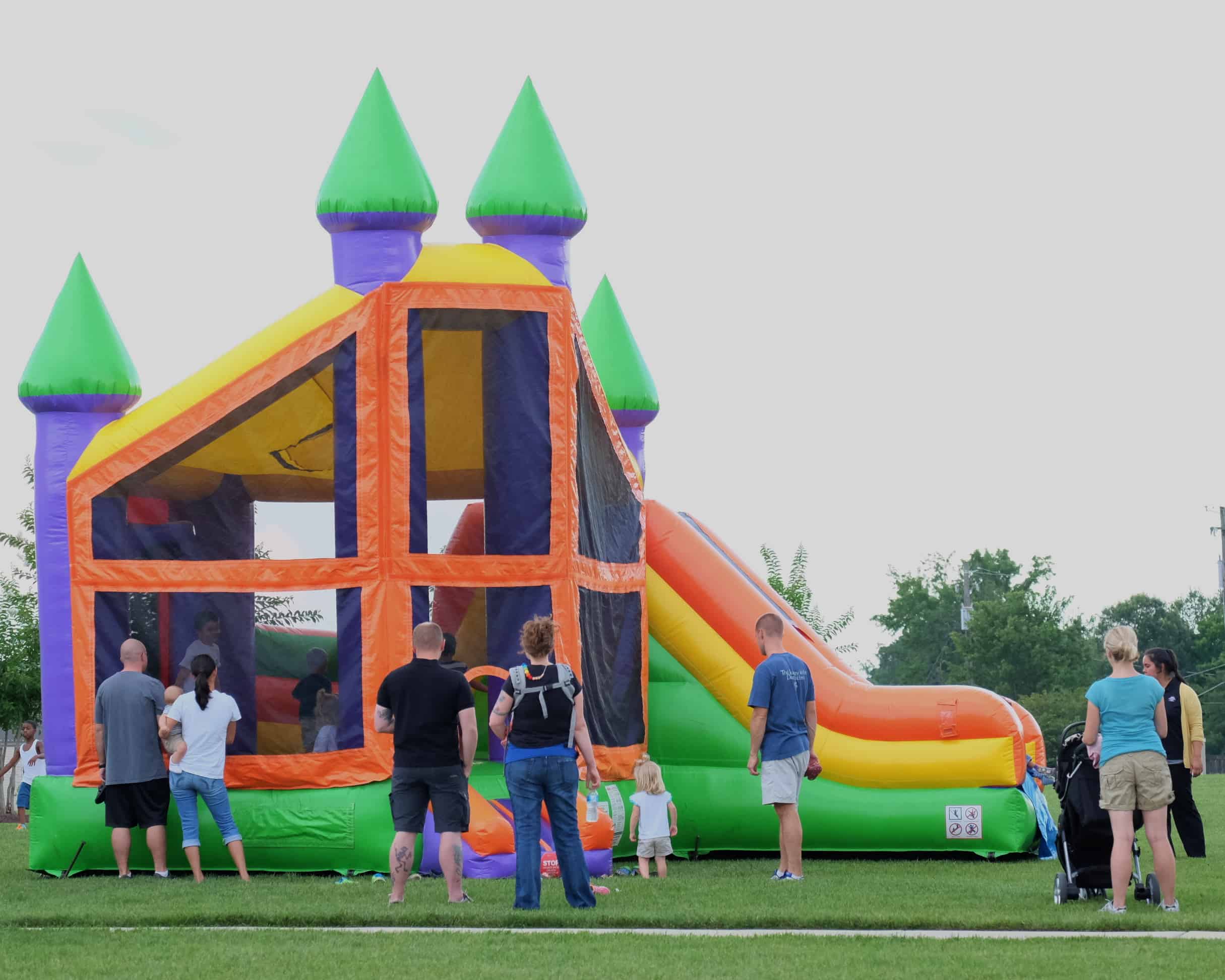Large colorful bounce house with attached slide set up on a grassy field with families and children gathered around for an outdoor event